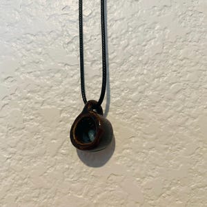 May include: A dark brown ceramic pendant necklace. The pendant is a small, hollow, cup-shaped design with a dark interior and a black cord. The necklace is displayed against a textured white background.