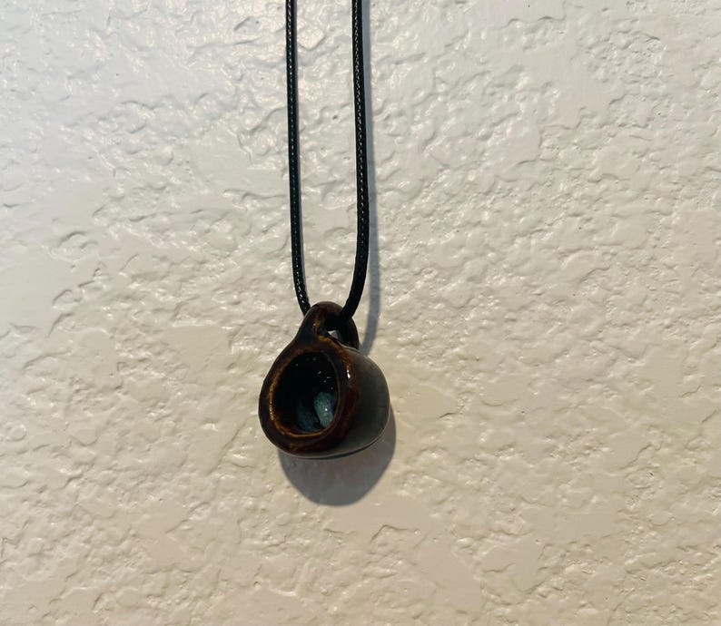 May include: A dark brown ceramic pendant necklace. The pendant is a small, hollow, cup-shaped design with a dark interior and a black cord. The necklace is displayed against a textured white background.