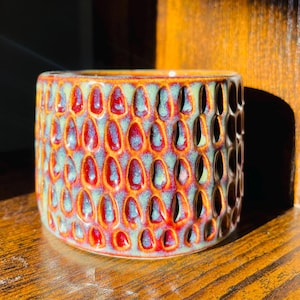 May include: A cylindrical ceramic planter featuring a repeating embossed teardrop pattern across its exterior. The surface displays a reactive glaze with deep red and orange tones transitioning into soft sage green highlights within the textured indentations.