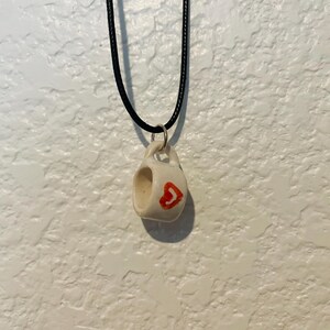 May include: A small, white ceramic mug pendant with a red heart design, suspended from a black cord necklace. The pendant is approximately 0.75 inches in height and width, with a small handle. The background is a textured white wall.