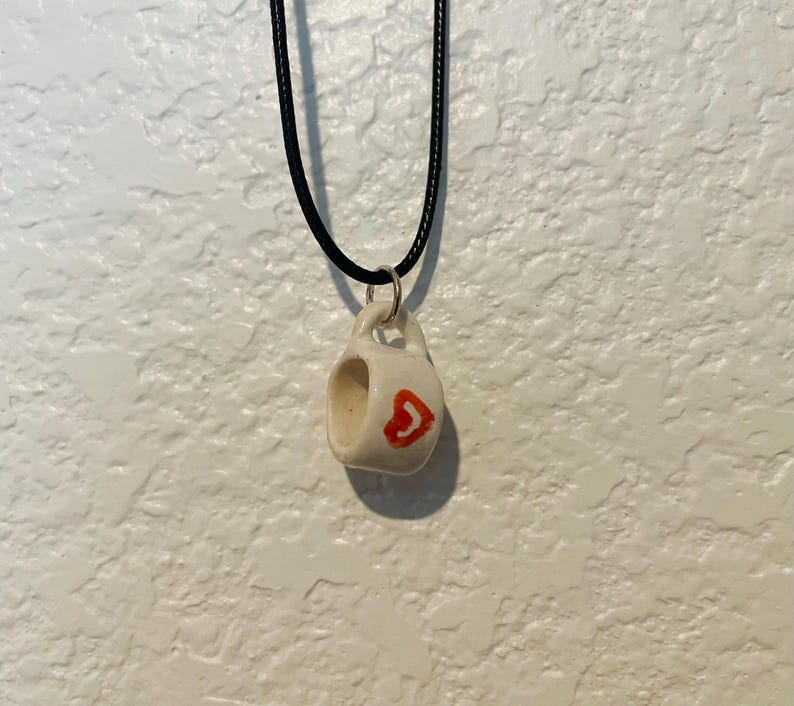 May include: A small, white ceramic mug pendant with a red heart design, suspended from a black cord necklace. The pendant is approximately 0.75 inches in height and width, with a small handle. The background is a textured white wall.