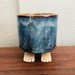 May include: A small, cylindrical ceramic planter with a blue glaze and brown rim. The planter is supported by two small, white, foot-shaped legs. The blue glaze has a mottled appearance, adding visual interest to the piece. This unique planter is suitable for small plants.