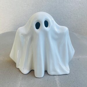 May include: A white ceramic ghost figurine with a glossy finish. The ghost has two black eye holes and a draped sheet-like form. The figurine is a decorative item, perfect for Halloween or spooky-themed decor.