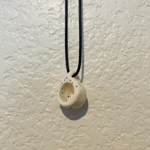 May include: A small, white ceramic pendant necklace with a black cord. The pendant is shaped like a miniature cup with a hole in the center and is speckled with blue, green, and yellow dots. The necklace hangs against a textured white background.