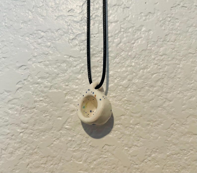 May include: A small, white ceramic pendant necklace with a black cord. The pendant is shaped like a miniature cup with a hole in the center and is speckled with blue, green, and yellow dots. The necklace hangs against a textured white background.