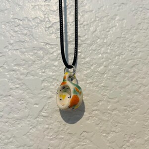 May include: A ceramic pendant necklace with a black cord. The pendant is white with colorful speckled accents of orange, yellow, and green. The pendant has a unique, curved shape with a small loop for the cord to pass through.