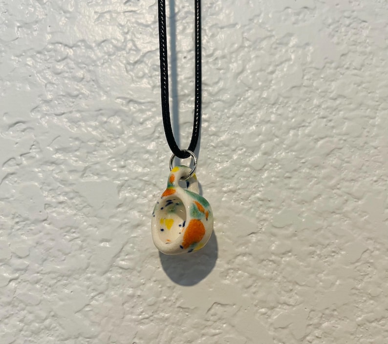May include: A ceramic pendant necklace with a black cord. The pendant is white with colorful speckled accents of orange, yellow, and green. The pendant has a unique, curved shape with a small loop for the cord to pass through.