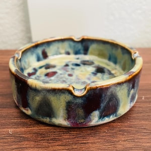 May include: A round ceramic ashtray with a glazed finish. The ashtray features a mix of colors, including blue, green, and maroon. The rim has a light brown color. The ashtray has three notches for resting cigarettes.