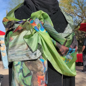 May include: A green silk scarf with a colorful abstract design is draped over a black sweater. The scarf features shades of green, blue, red, and brown. The scarf is wrapped around the neck and hangs down the front.