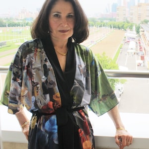 May include: A woman wearing a colorful kimono-style robe with a black sash. The robe features a painting-like design with figures and a green landscape. She is standing on a balcony with a city and race track in the background.