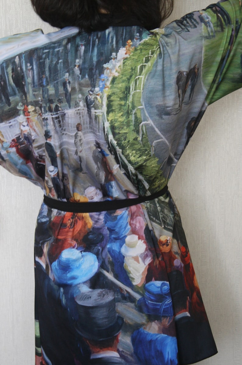 May include: A kimono-style garment featuring a vibrant, impressionistic painting of a horse race scene. The artwork depicts spectators in hats and suits, with a racetrack and horses visible. The garment is cinched at the waist with a black belt.