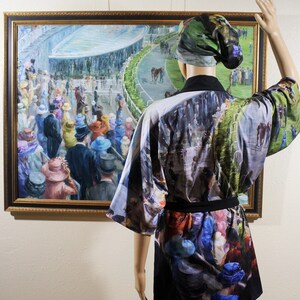 May include: A mannequin displays a kimono-style robe and matching head covering, both featuring a print of a horse race scene. The robe is black with a colorful print of people and horses. A framed painting of a horse race is in the background.
