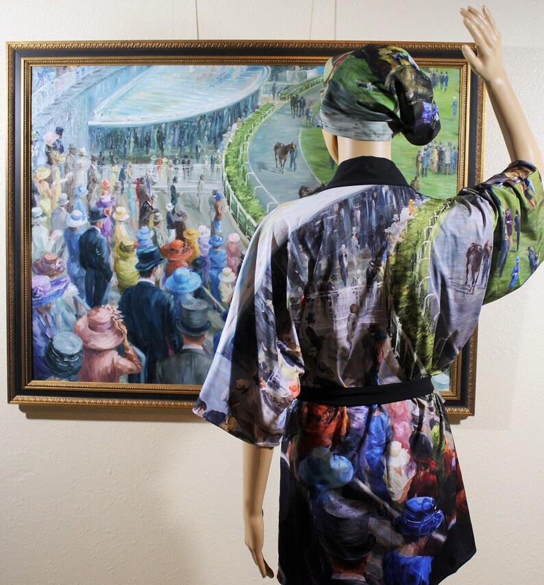 May include: A mannequin displays a kimono-style robe and matching head covering, both featuring a print of a horse race scene. The robe is black with a colorful print of people and horses. A framed painting of a horse race is in the background.