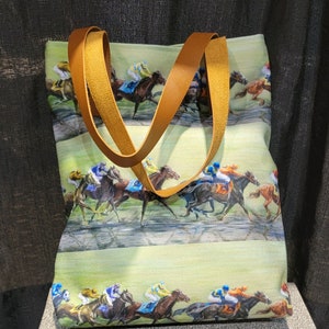 May include: A tote bag featuring a horse racing scene with jockeys in colorful silks. The bag has a light green background and brown leather handles. The image depicts horses in mid-race, with a focus on speed and competition.