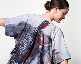 Puede incluir: Una blusa holgada de color gris claro con una pintura detallada de una figura a caballo, sobre un fondo montañoso. La obra de arte incluye tonos de rojo, azul y gris, creando una prenda única y artística.
