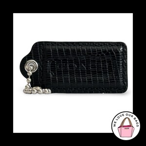 May include: A black rectangular leather bag tag with a textured lizard-skin pattern and a silver-toned metal ball chain. The word COACH is embossed in the center of the tag. A circular logo in the corner features a pink tote bag and the text WE LOVE OUR BAGS.