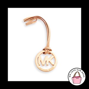 May include: A rose gold-toned metal Michael Kors circular logo charm suspended from a light tan leather strap. The circular charm features the letters MK in the center with the brand name Michael Kors embossed along the bottom edge of the ring.