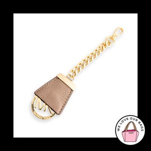May include: A gold-toned metal chain keychain featuring a taupe-colored textured leather tab and a circular gold-toned metal charm with the letters MK and the text MICHAEL KORS engraved on the front. The keychain includes a lobster claw clasp.