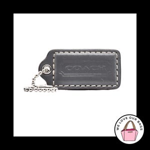 May include: A dark gray leather Coach luggage tag with white stitching and a silver ball chain. The Coach logo is embossed on the tag. The tag is rectangular with a rounded top and a small loop for the chain.