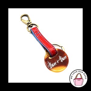 May include: A red leather keychain with a light blue backing and gold-colored hardware. The keychain has a round, tortoiseshell-patterned charm with the text "Barr + Barr" in white script. A small, pink bag logo is in the bottom right corner.
