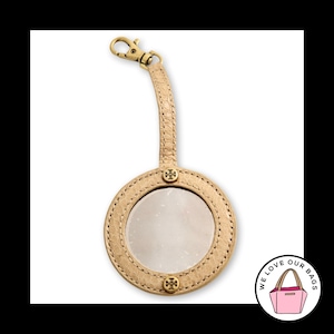May include: A beige textured leather circular luggage tag with a clear window, gold-tone metal hardware, a lobster clasp strap, and two small gold-tone metal logos at the top and bottom of the circular frame, featuring a light beige finish.