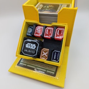 以下が含まれることがあります： 黄色いカードゲーム収納ボックス。複数のコンパートメントがあり、カードの束と、ラベルの付いた小さなゲームピースがいくつか見えます。「STAR WARS UNLIMITED」の文字がゲームピースに表示されています。