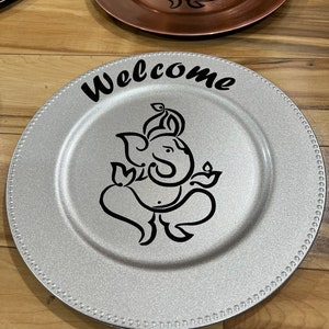 May include: Silver charger plate with a black and white design of a Hindu deity, Ganesha, and the word "Welcome" in black lettering.