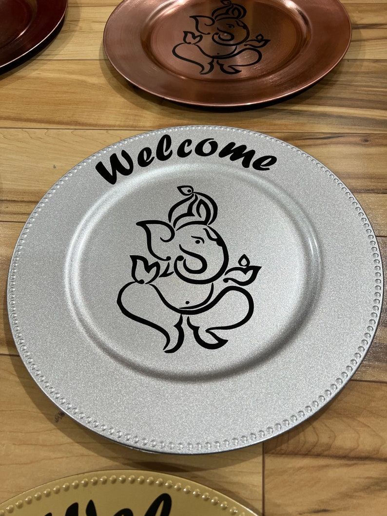 May include: Silver charger plate with a black and white design of a Hindu deity, Ganesha, and the word "Welcome" in black lettering.