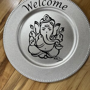 May include: Silver charger plate with a black and white image of Ganesha and the word "Welcome" in black lettering.