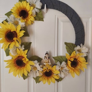 Puede incluir: Una corona de tela gris decorada con girasoles amarillos y margaritas blancas. Una peque&ntilde;a figura de una persona est&aacute; en el centro de la corona.