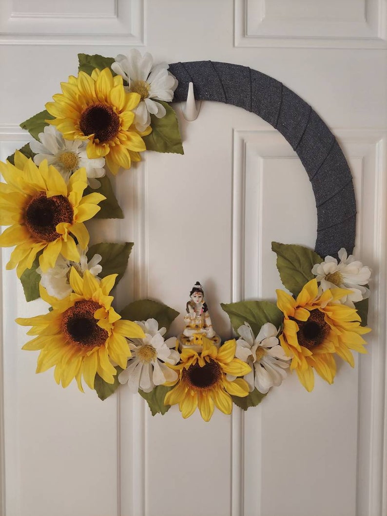Puede incluir: Una corona de tela gris decorada con girasoles amarillos y margaritas blancas. Una peque&ntilde;a figura de una persona est&aacute; en el centro de la corona.
