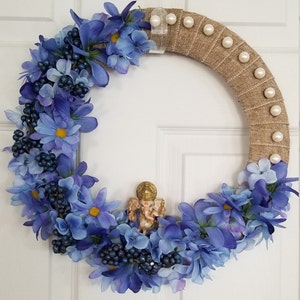 Puede incluir: Una corona floral azul y blanca con una peque&ntilde;a estatua dorada de Ganesha en el centro. La corona est&aacute; hecha de arpillera y tiene detalles de perlas blancas.