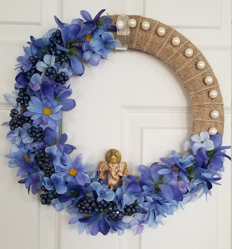 Puede incluir: Una corona floral azul y blanca con una peque&ntilde;a estatua dorada de Ganesha en el centro. La corona est&aacute; hecha de arpillera y tiene detalles de perlas blancas.