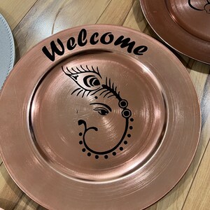 May include: A rose gold charger plate with a black design of an elephant with a feather and the word "Welcome" on the top.