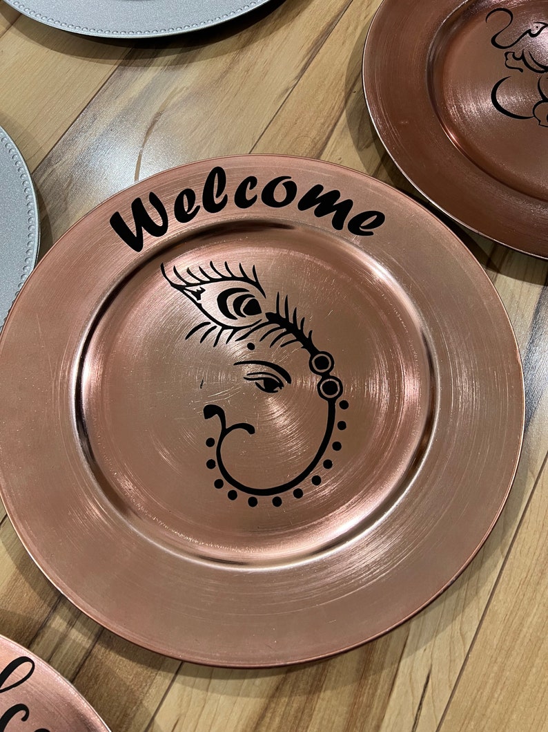 May include: A rose gold charger plate with a black design of an elephant with a feather and the word "Welcome" on the top.