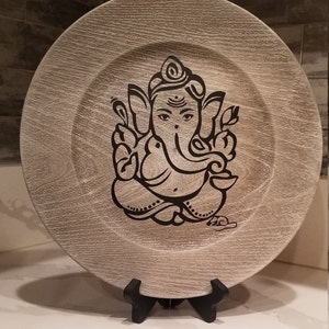 May include: A decorative wooden plate with a black and white image of the Hindu deity Ganesha. The plate is on a black stand.