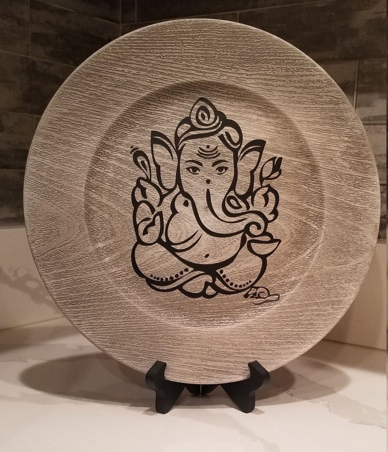 May include: A decorative wooden plate with a black and white image of the Hindu deity Ganesha. The plate is on a black stand.