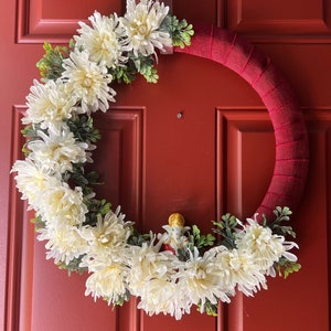 Puede incluir: Una corona de flores blancas con hojas verdes en una puerta roja. Una peque&ntilde;a figura est&aacute; en el centro de la corona.