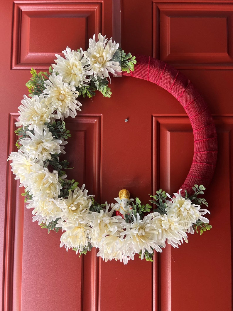 Puede incluir: Una corona de flores blancas con hojas verdes en una puerta roja. Una peque&ntilde;a figura est&aacute; en el centro de la corona.