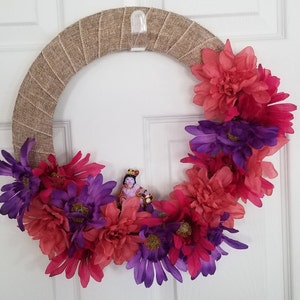 Puede incluir: Una corona de arpillera decorada con flores rosas y moradas. Una peque&ntilde;a figura de una persona con una corona y ropa morada est&aacute; en el centro de la corona.