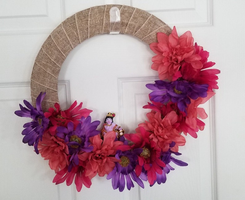 Puede incluir: Una corona de arpillera decorada con flores rosas y moradas. Una peque&ntilde;a figura de una persona con una corona y ropa morada est&aacute; en el centro de la corona.