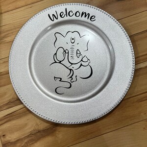 May include: A silver charger plate with a beaded edge features a black and white illustration of Ganesha, the Hindu god of beginnings, and the word "Welcome" in black lettering.