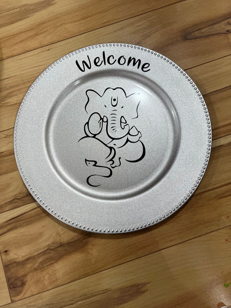 May include: A silver charger plate with a beaded edge features a black and white illustration of Ganesha, the Hindu god of beginnings, and the word "Welcome" in black lettering.