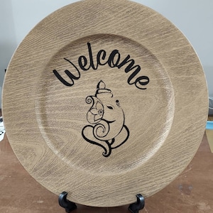 May include: A round, wooden charger plate with a woodgrain pattern. The plate has a black design of an elephant with the word "Welcome" written above it.