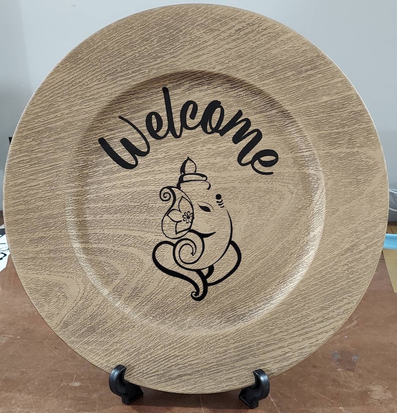 May include: A round, wooden charger plate with a woodgrain pattern. The plate has a black design of an elephant with the word "Welcome" written above it.