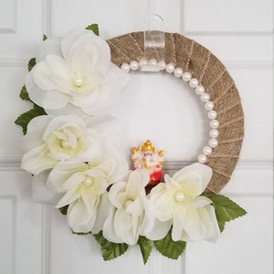Puede incluir: Una corona de arpillera marr&oacute;n decorada con flores de tela blanca y una guirnalda de perlas. Una peque&ntilde;a figura de la deidad hind&uacute; Ganesha se coloca en el centro de la corona.