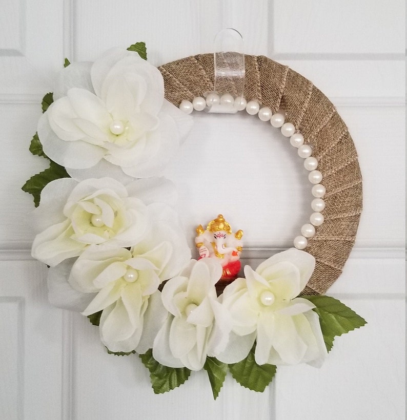 Puede incluir: Una corona de arpillera marr&oacute;n decorada con flores de tela blanca y una guirnalda de perlas. Una peque&ntilde;a figura de la deidad hind&uacute; Ganesha se coloca en el centro de la corona.