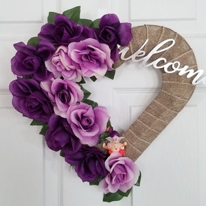 Puede incluir: Una corona en forma de coraz&oacute;n hecha de arpillera con rosas artificiales moradas y lavanda. La corona tiene un letrero de "Bienvenido" plateado adjunto.