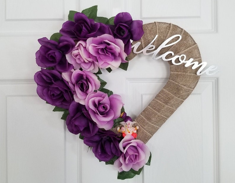 Puede incluir: Una corona en forma de coraz&oacute;n hecha de arpillera con rosas artificiales moradas y lavanda. La corona tiene un letrero de "Bienvenido" plateado adjunto.