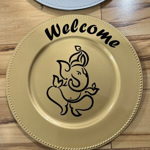 May include: Gold charger plate with a black outline of Ganesha and the word "Welcome" in black cursive lettering.